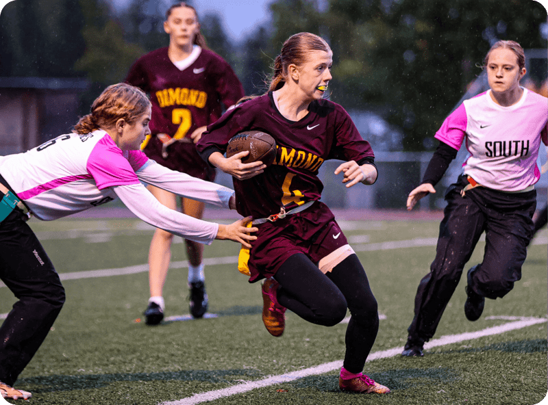 A female flag football player in a maroon Dimond jersey tucks the ball and runs as two South defenders close in